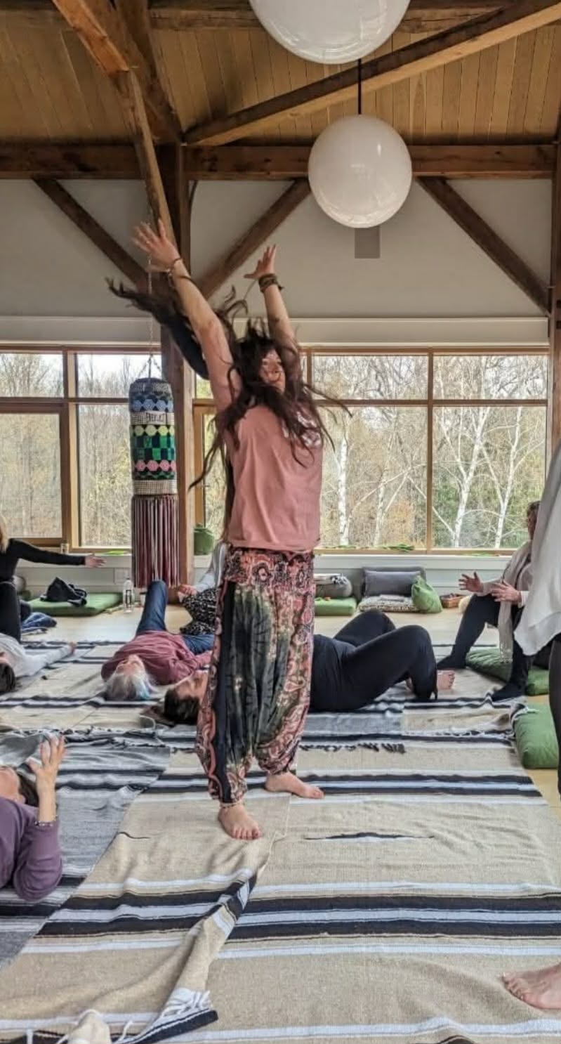 Melisa standing with both arms raised, leading a group breathwork session in a timber-framed room while participants lie on blankets on the floor.